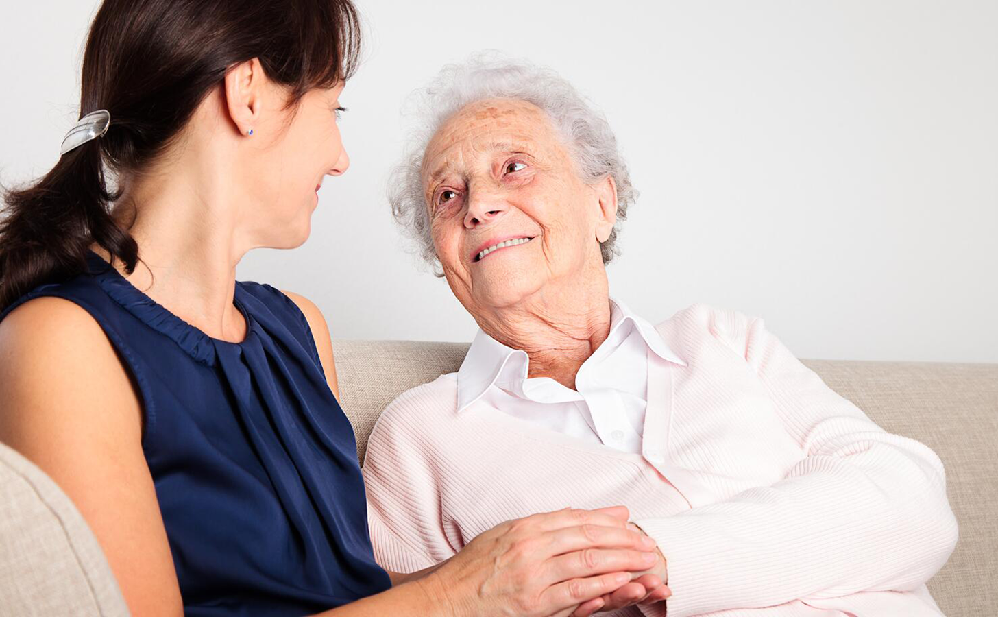 A female caregiver sits with an elderly woman while holding her hand and looking at her.