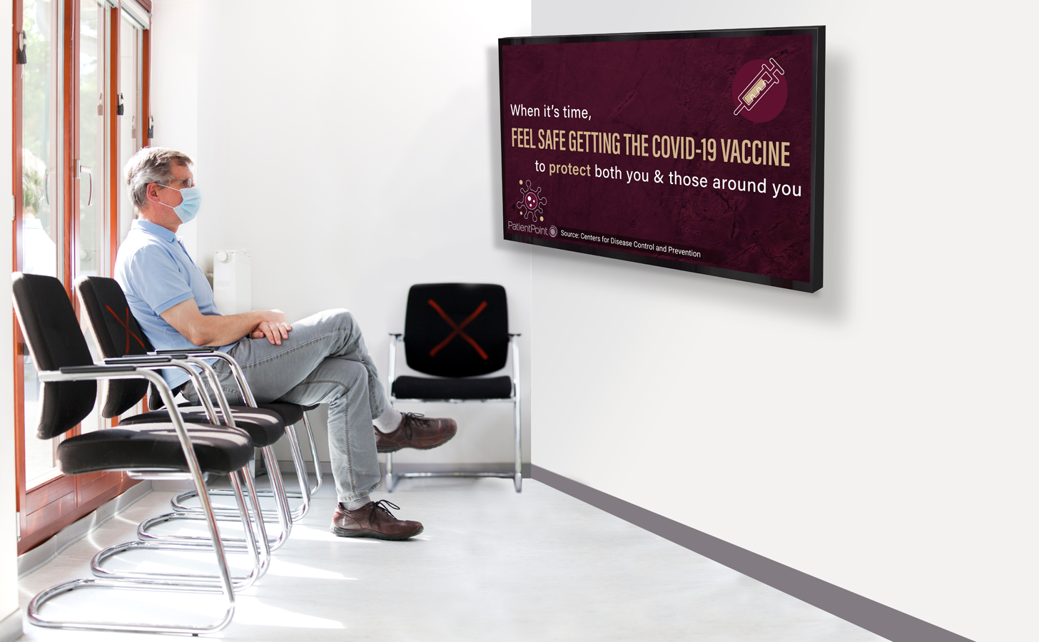 A middle-aged man wearing a mask sits in the waiting room of his physician's office and watches content on a TV screen about the safety of the COVID-19 vaccine.