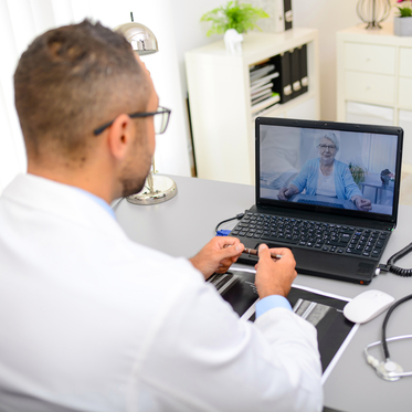 Male doctor sitting at desk looking at laptop showing virtual health consultation with older female patient