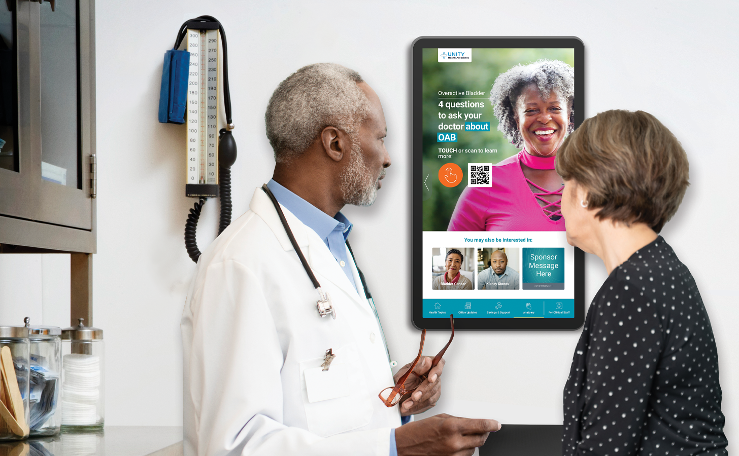 A mature male physician reviews content about overactive bladder on a wall-mounted touchscreen in an exam room with a middle-aged female patient. 
