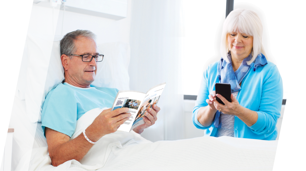 Male patient in hospital bed reading patient handbook with female caregiver standing next to him looking at her phone.