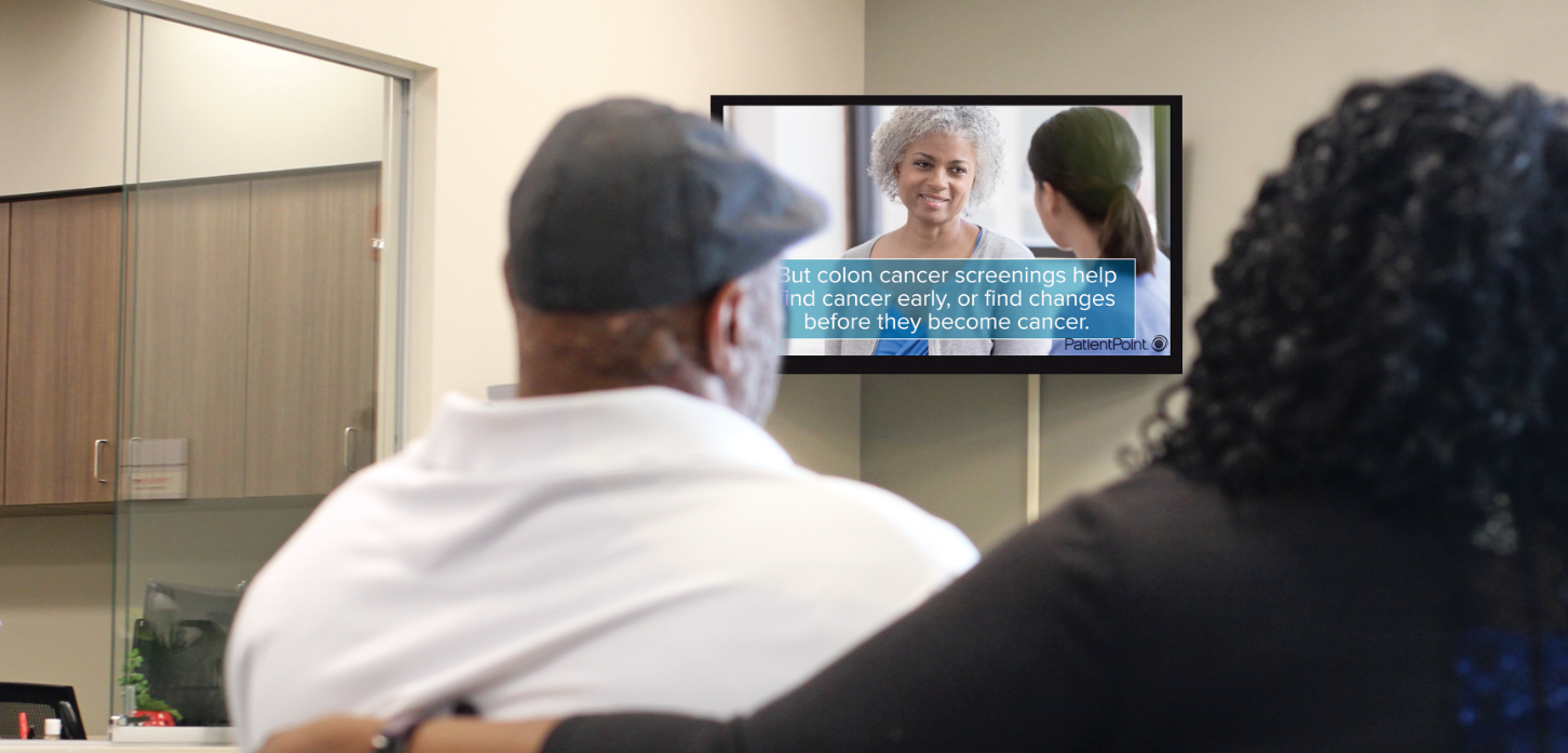 Couple watching waiting room screen with colon cancer screening info