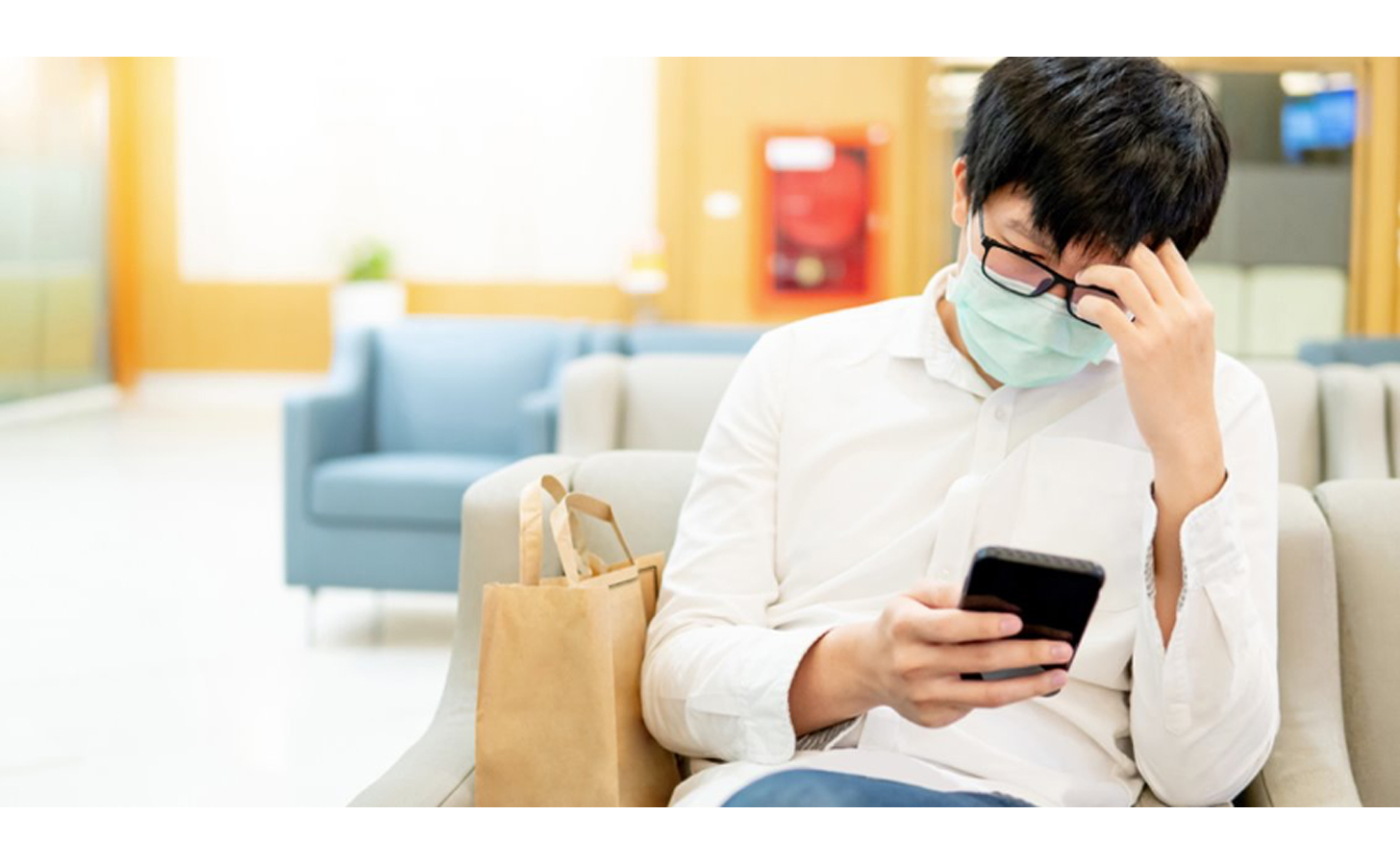 A young asian man sits in the waiting room wearing a mask while looking at his smartphone.