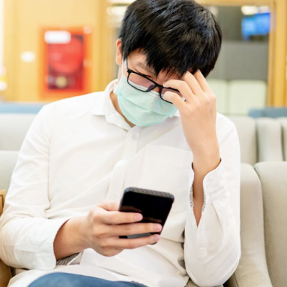 Middle-aged Asian man wearing facemask sitting in doctor's office waiting room looking at mobile phone.
