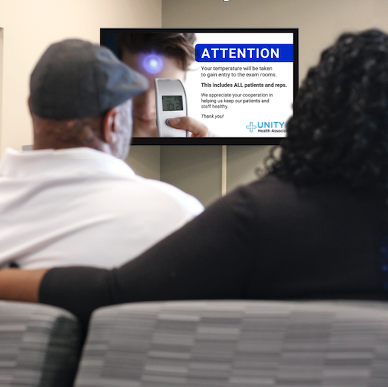 Patient and caregiver in waiting room watching PatientPoint Communicate screens with Covid-19 custom messages.