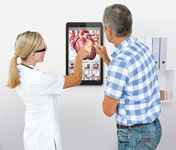 Woman doctor and male patient explore a digital anatomical of the heart on an interactive exam room touchscreen.